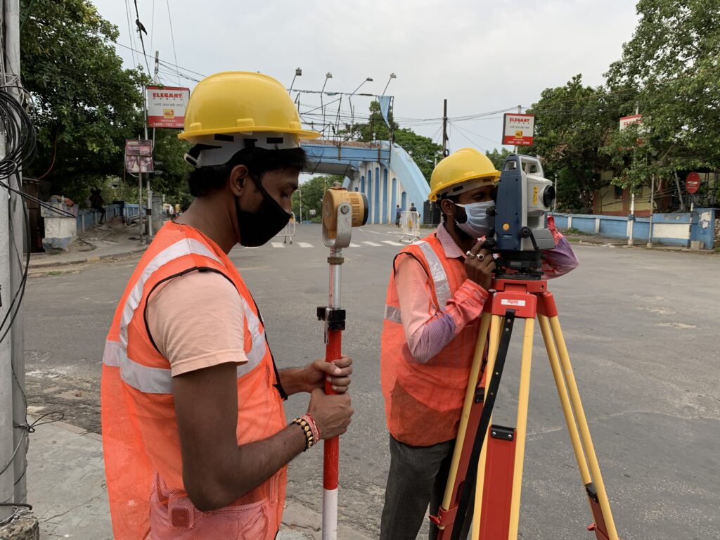 Two survey workers in yellow hard hats and orange high-visibility vests using a total station on a street.