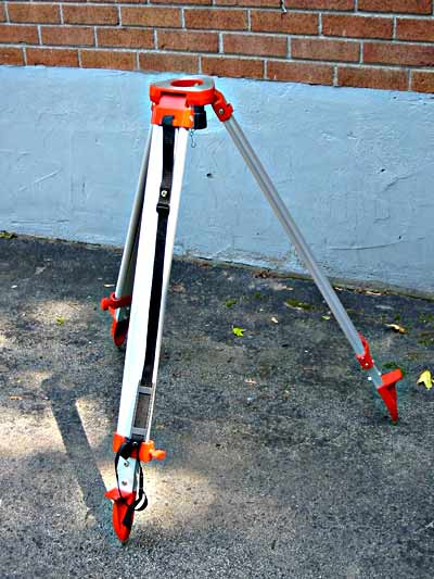 Silver surveyor’s tripod with red fittings and pointed feet, standing beside a brick wall.