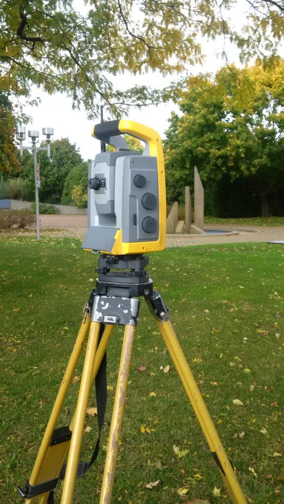 Yellow-and-gray robotic total station on a yellow tripod outdoors beside grass and trees.